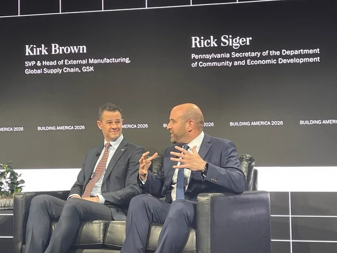 Secretary Rick Siger and GSK SVP in business suits seated on stage during a Building America 2026 event, engaged in conversation with a black backdrop displaying their names and titles.