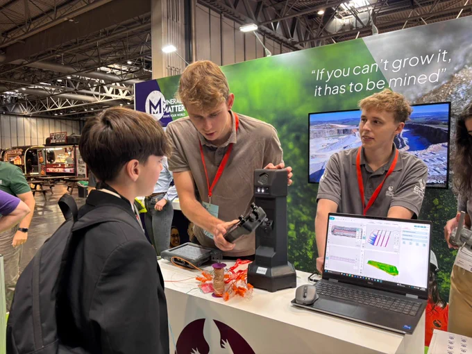 An exhibition booth features representatives engaging with a visitor. A person is demonstrating equipment on a stand with colorful wrappers nearby. Another representative is standing by a laptop displaying data. The backdrop includes a logo and motto related to mining.