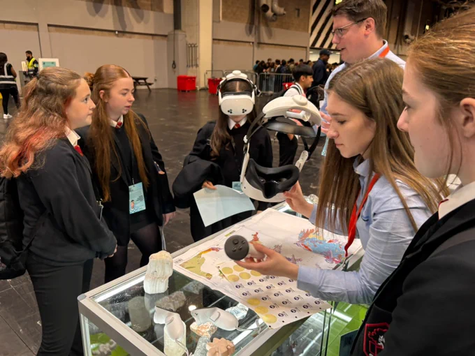 A group of people are gathered around an exhibit table displaying various scientific models and a virtual reality headset. One person is explaining the items, while others listen attentively. The setting appears to be a busy exhibition or event space.