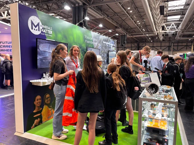 A group of people are gathered near the "Minerals Matter" booth at an exhibition. Some are interacting, and others are observing the display. The booth features a screen displaying the booth's logo, a green-themed backdrop, and a glass case with mineral samples. The area is busy, with many attendees walking around.