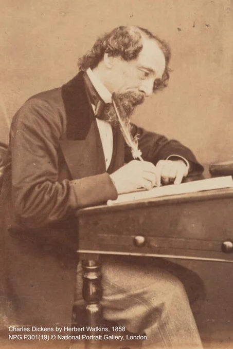A sepia photograph of Charles Dickens, in Victorian dress, seated at a desk as he writes