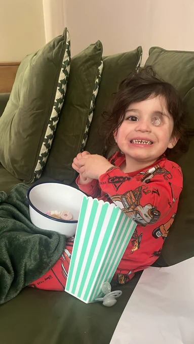 Nuala sits on the sofa, with a bag of popcorn and bowl of party rings on her lap. She waers red pyjamas