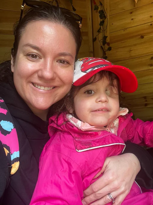 Megan is a white woman with brunette hair. She has nuala on her lap, wearing a pink raincoat and cap. Nuala has eye loss.
