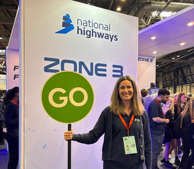 A person holding a green "Go" sign stands in front of a display with the "National Highways" logo and "Zone 3" text. The individual wears a badge, and the setting appears to be a busy event or exhibition space.