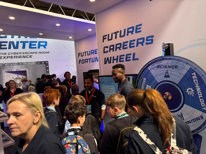 A group of people are gathered around a booth featuring a "Future Careers Wheel" at a technology event. The wheel has sections labeled "Science" and "Technology." Banners with text about future careers and fortune are displayed. People are engaged in conversation, and the area is well-lit and busy.