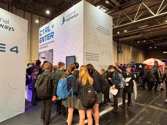 A group of individuals stand in line at a National Highways booth inside a large indoor exhibition space. The booth features a tall, white panel with text related to digital solutions.