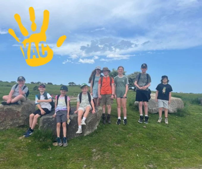 Nine young people wearing hats and backpacks stand around large rocks in the middle of a grassy field on a sunny day. 