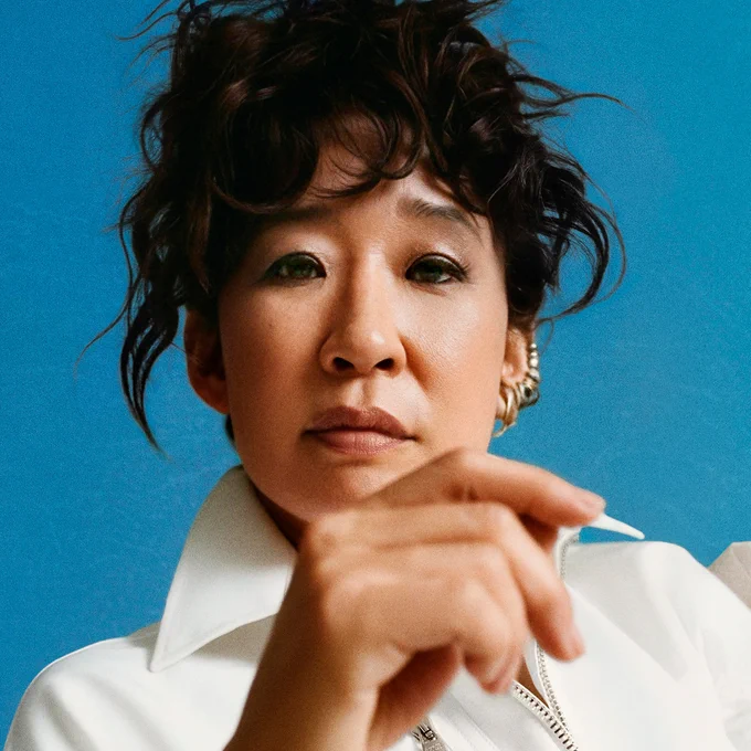 Sandra Oh wears a white jacket and has her hair up. The background behind her is bright blue.