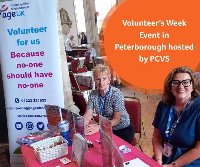 Photograph of Carol and Anita sat down at a table in a church hall. They are looking and smiling at the camera. On the table is a collection of leaflets and marketing materials they also have a pull up banner to the left of them. The banner Says Volunteer for us, because no one should have no one’. To the right corner is an orange circle with text that says ‘Volunteer’s Week Event in Peterborough hosted by PCVS’