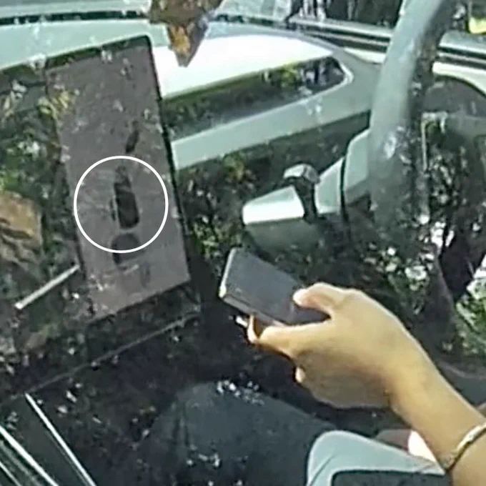 A still frame from a video showing the simulated overhead view displayed on a Tesla infotainment screen, the driver is interacting with a handheld mobile telephone, this is illegal even when stationary in the United Kingdom. The driver can expect to receive points on her licence.