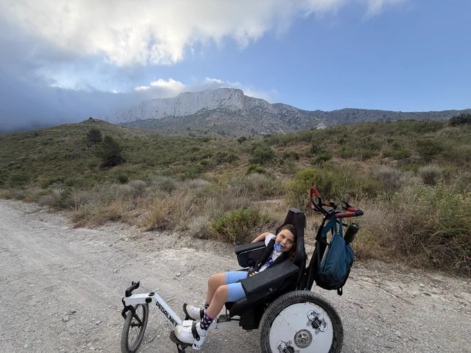 julia en su silla de correr con el mordedor en la boca y el tajo de peña blanca al fondo rodeado de nubes (es una pared enorme vertical de color blanco)