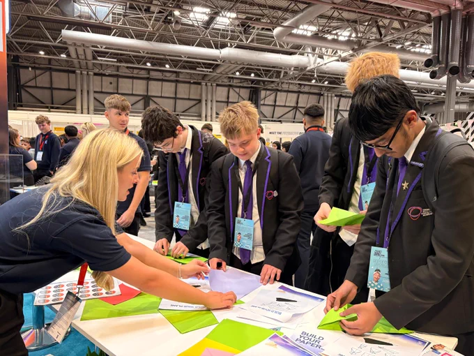 A group of students in school uniforms are gathered around a table, engaging in a hands-on activity involving colorful paper. An instructor is assisting them. The setting is an indoor exhibition or event space with high ceilings and displays.
