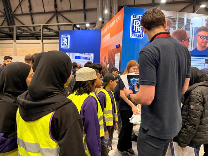 A group of young students wearing safety vests stands in front of a Rolls‑Royce booth at an indoor event. An adult holding a tablet engages with the group. The setting is a busy exhibition hall.