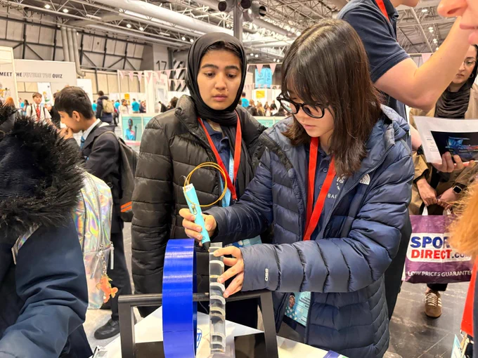 Two individuals in a busy indoor setting are engaged in an activity involving a structure with blue and black components. One person is attentively working on the structure, holding a pen-like object. The other observes closely. In the background, there are people, displays.