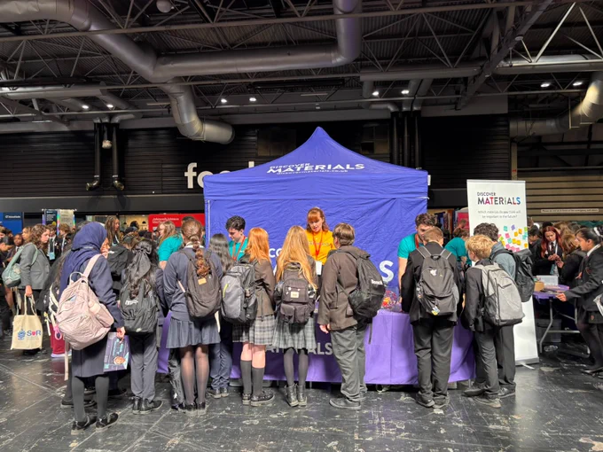 Crowd of people stand in front of a booth with a purple tent labeled "DISCOVER MATERIALS." They appear to be engaging with representatives behind the booth. Indoors at an event with other booths visible in the background.