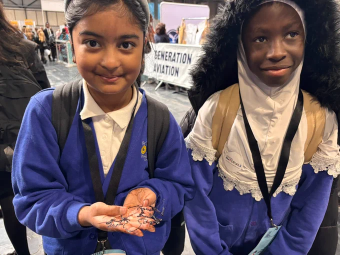 Two children in blue uniforms are smiling at an indoor event. One holds a small wire model of an atom.