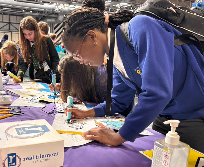 Students are engaged in an activity using 3D printing pens at a table covered with a purple cloth. Other items like color pens and a bottle of hand sanitizer are on the table. The setting is a busy indoor event.