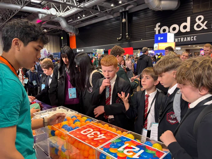 A group of young people in school uniforms are gathered around a table filled with colorful balls, listening intently to a person in a teal shirt who is explaining something. The atmosphere is lively and focused.