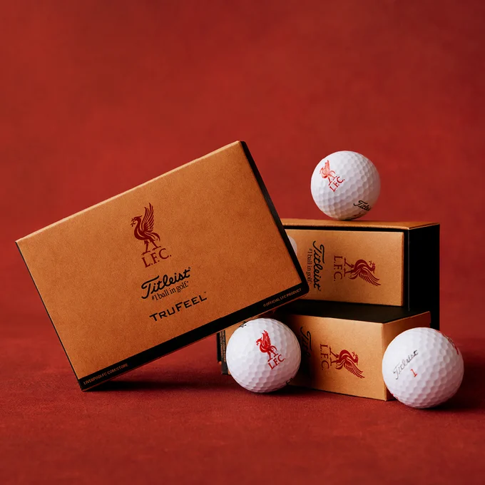 Liverpool FC and Titleist TruFeel golf balls and branded packaging arranged on a red surface, featuring gold boxes with the LFC crest and white balls with printed logos.