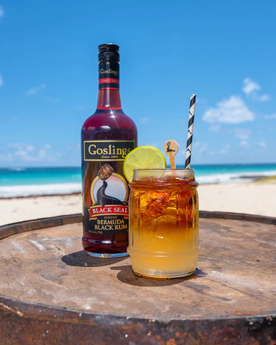 A bottle of Gosling’s Black Seal Bermuda Black Rum and a freshly poured Dark 'n Stormy® cocktail sitting on a rustic wooden barrel on a pink sand beach. The layered drink is garnished with a fresh lime wedge and a black-and-white striped straw, with the turquoise ocean and blue sky in the background, celebrating International Dark 'n Stormy Day in Bermuda.