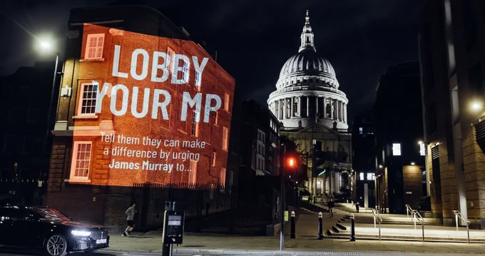 A message in white on a red background saying, 'Lobby Your MP,  Tell them they can make a difference by urging James Murray to ...' projected on a wall with S Paul's Cathedral at night in the background.