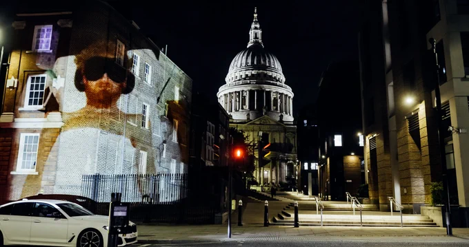 A photograph of a young man with red/brown hair wears an eye mask and headphones lying still in bed is projected on a wall with S Paul's Cathedral at night in the background.