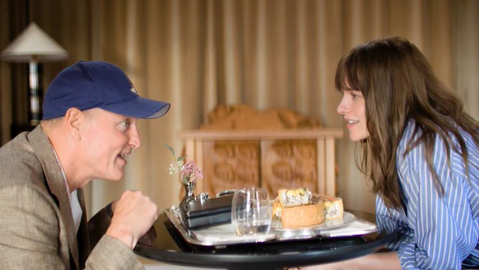 Woody Harrelson and Kristen Stewart as dad and daughter in Full Phil. A man in a baseball cap sits opposite a woman in a striped shirt, there is a tray of food between them