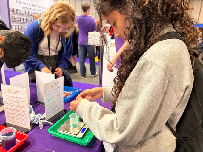 A person in a gray sweatshirt engages in a hands-on science activity at a table. Nearby, others focus on their own tasks. The environment is a science fair or educational event, with informational posters and purple tablecloths visible.