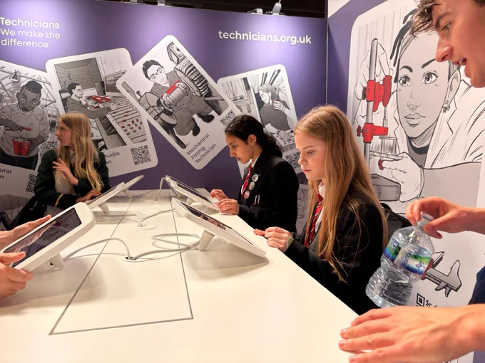 A group of individuals is engaged with tablets on a white table. The backdrop features illustrations of technicians, tools, and the logo “technicians.org.uk.” One person is holding a water bottle. The setting appears to be centered around promoting the role of technicians.