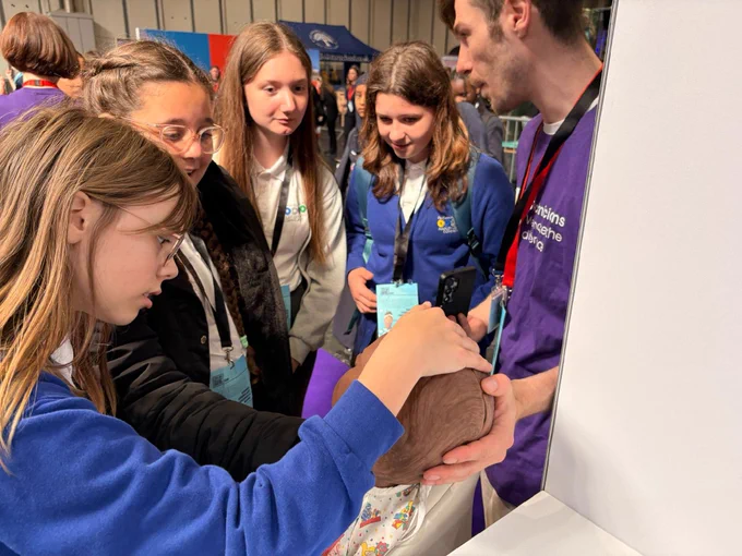 A group of individuals is gathered around an exhibit at what appears to be a science fair or educational event. They are observing a brown object that one person is holding. The person holding the object is wearing a purple shirt with text on it. Everyone looks attentive and engaged. The setting is indoors with informational booths visible in the background.