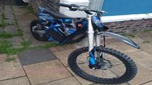 A blue and black electric dirt bike with distinctive graphics, parked on a paved surface.