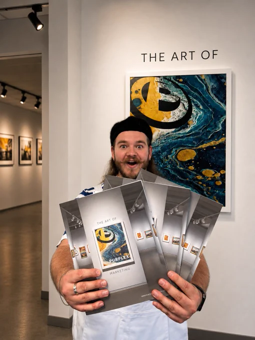 A man excitedly holds multiple copies of a book titled 'The Art of Marketing' in an art gallery. The mood is lively and engaging.