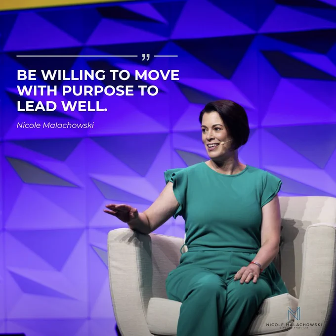 Nicole Malachowski sits on stage in a light-colored chair, wearing a teal outfit and a headset microphone, gesturing as she speaks against a purple geometric backdrop. Large overlaid text reads: “BE WILLING TO MOVE WITH PURPOSE TO LEAD WELL.” The quote is attributed to Nicole Malachowski. The Nicole Malachowski & Associates logo appears in the bottom-right corner.