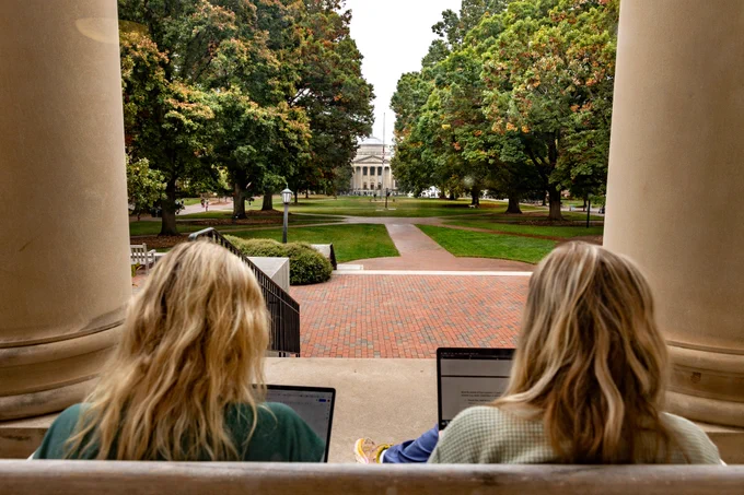 Two female students are working on their respective laptops on the steps of South Building looking at Wilson Library. 