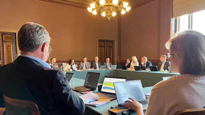 Pyöreän pöydän keskustelutilaisuus ministeriön kokoushuoneessa: useita osallistujia istuu suuren pöydän äärellä läppärit ja muistiinpanovälineet edessään, ja tilassa on korkea katto sekä kattokruunu.
