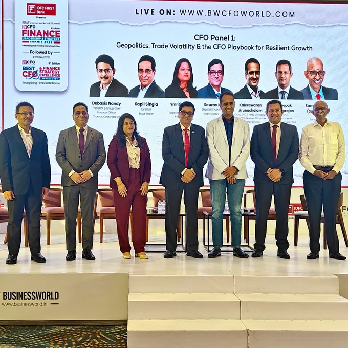 Seven professionally dressed panelists stand on stage in front of a large screen displaying event and speaker information at a finance conference.