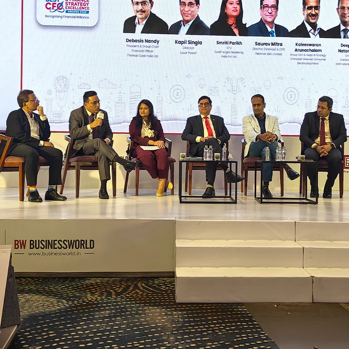 Six panelists sit on stage during a business conference, engaged in discussion, with a projected event banner behind them displaying their names and photos.