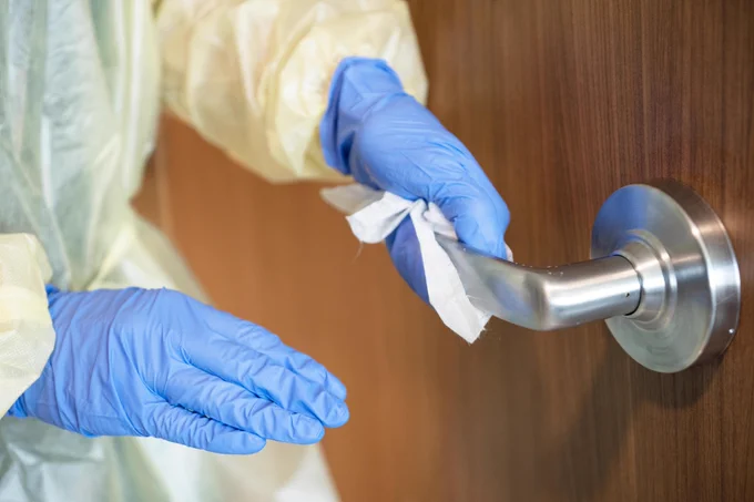 Obscured EVS staff wearing yellow disposable gown and blue gloves is wiping a door handle.