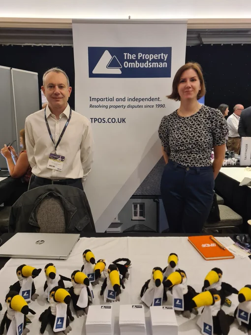 A man and woman in smart business clothing are stood in front of a roller banner promoting The Property Ombudsman (TPO). On the table in front of them are TPO-branded toy toucans.