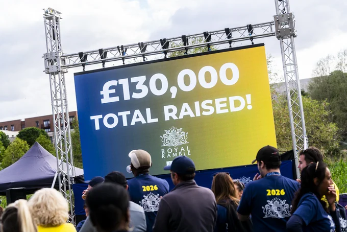 A large outdoor screen displays "£130,000 Total Raised!" with a "Royal Ramble" logo beneath. A crowd of people, some wearing blue shirts with "2026" printed on the back, stand in front of the screen. There are trees and a building in the background.