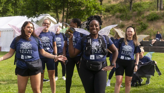 A group of people walking outdoors, wearing matching T-shirts with "Royal Ramble" printed on them. They have fairy wings on their backs and are smiling. There is a white tent and trees in the background.
