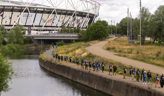A group of people, wearing matching shirts, are walking along a riverside path. In the background is the London Stadium. The area is surrounded by greenery, and the sky is overcast.