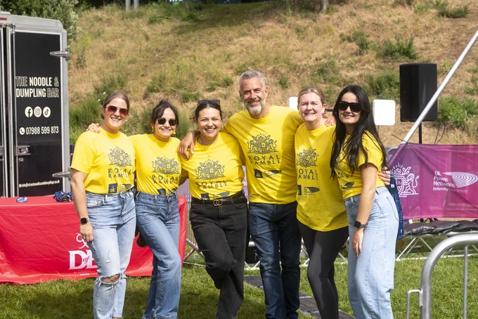 A group of people wearing matching yellow Royal Ramble t-shirts is posing together outdoors in a grassy area. They are smiling and standing in front of a mobile food truck and event banners.