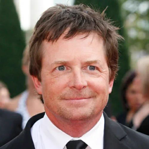 Michael J. Fox wears a black suit, white dress shirt, and black tie at a formal outdoor event. He faces the camera with a slight smile. His short brown hair is slightly tousled, and he has light stubble. Blurred people, tall greenery, and buildings are in the background.