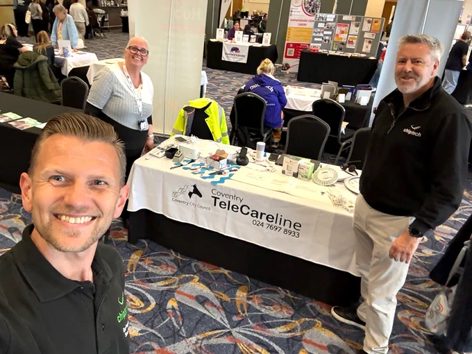 A Coventry TeleCareline booth at an indoor event with informational materials displayed on the table. On the left is a lady in a white top, and on the right is a man in a black Chiptech-branded top. Holding the camera in selfie mode is a man with a Chiptech-branded t-shirt on, smiling.