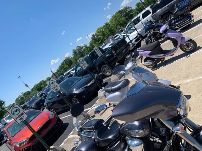 A parking lot with several parked motorcycles and scooters, including a purple scooter. Cars and trucks are parked in nearby spaces. Signs indicate designated motorcycle parking. Trees and a clear blue sky are in the background.