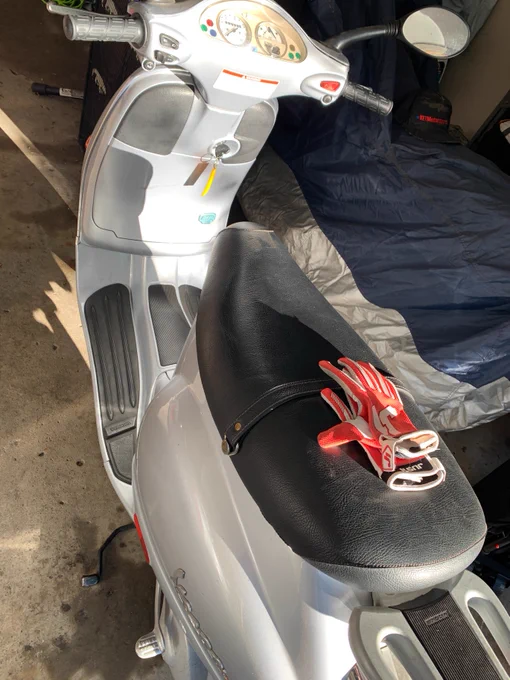 A white Vespa scooter with a black seat is parked indoors. Red gloves are placed on the seat. The scooter is next to a covered object, likely another vehicle.