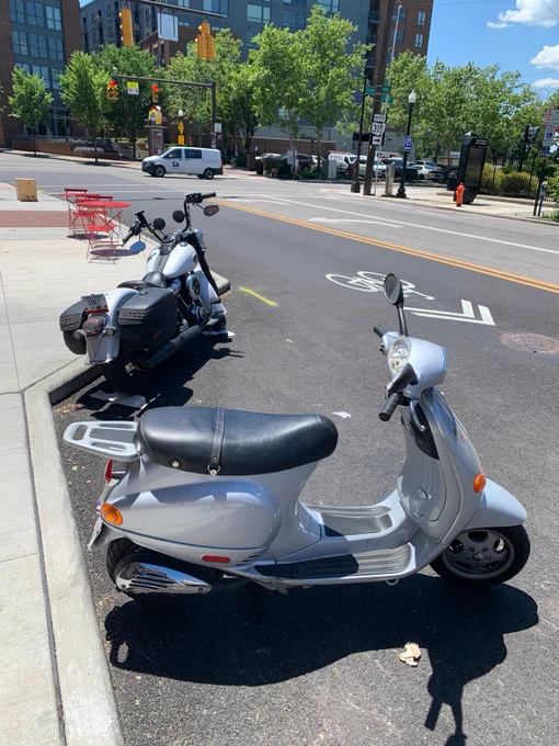 A silver scooter and a black Harley-Davidson motorcycle are parked on the side of an urban street. Red construction barriers and buildings with green trees are in the background. The street has clear bike lane markings.