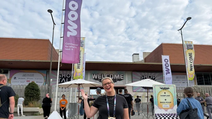 A person smiles and poses at the entrance of Shoptalk Europe next to flags that say "Woo: Commerce for what's next.”