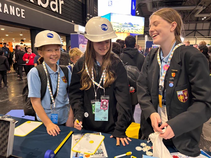 Three people wearing helmets are standing at an event booth. They are interacting with a table covered with various items, including papers and rulers. The event appears to be in a large conference hall with other attendees in the background.